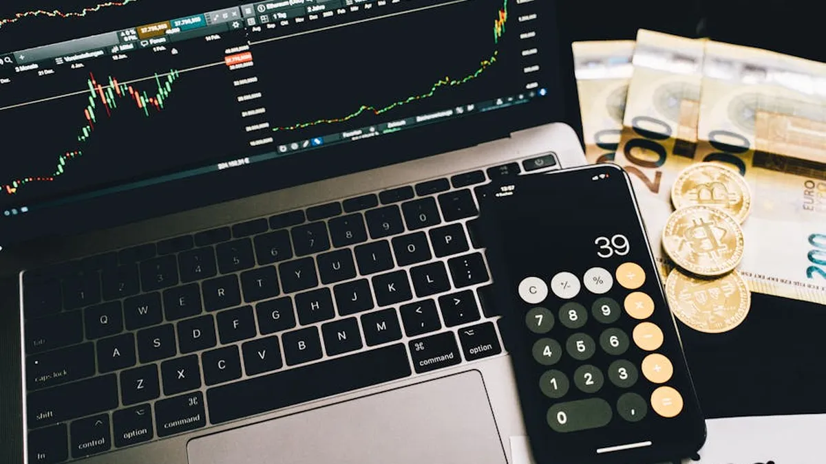 Laptop screen with trading charts, calculator app, Euros and Bitcoin coins on desk.