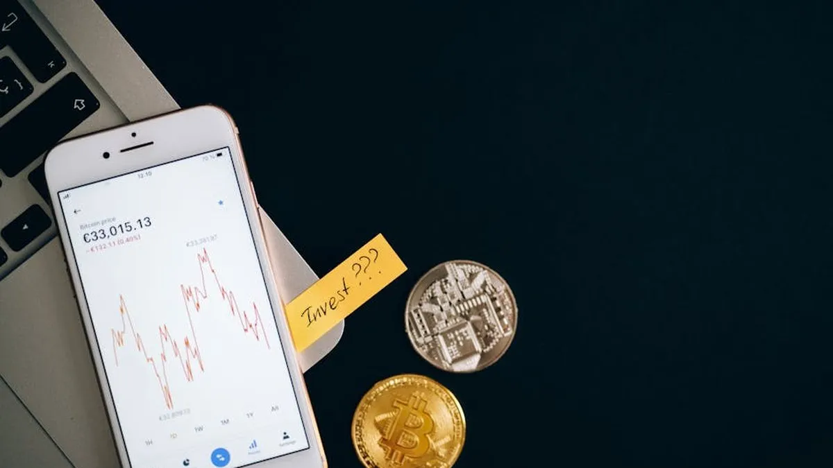 A smartphone displaying cryptocurrency prices beside coins and a laptop, symbolizing investment.